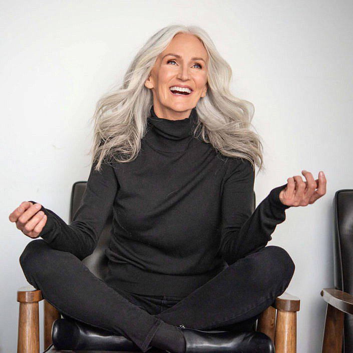 A woman with long, wavy gray hair, wearing an all-black outfit white sitting on the chair